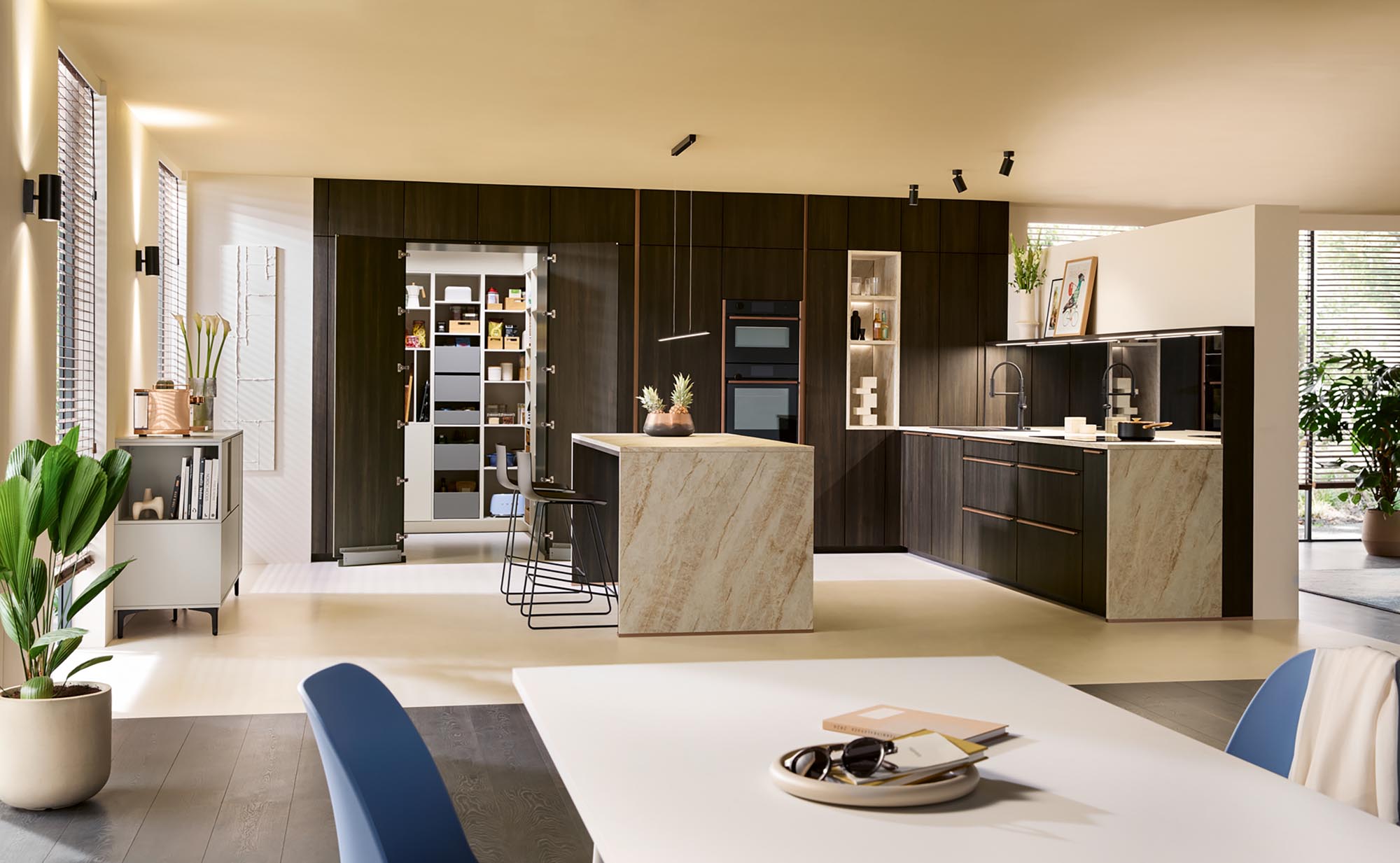 Moderne Küche mit dunklen Holzschränken, einer großen Kochinsel aus beigem Marmor, Einbaubacköfen und einer geräumigen begehbaren Speisekammer, die vom Schüller-Hauswirtschaftsraum inspiriert ist. Natürliche Lichtstrahlen durchfluten den Raum und bringen die Pflanzen sowie einen weißen Tisch mit blauen Stühlen besonders gut zur Geltung.